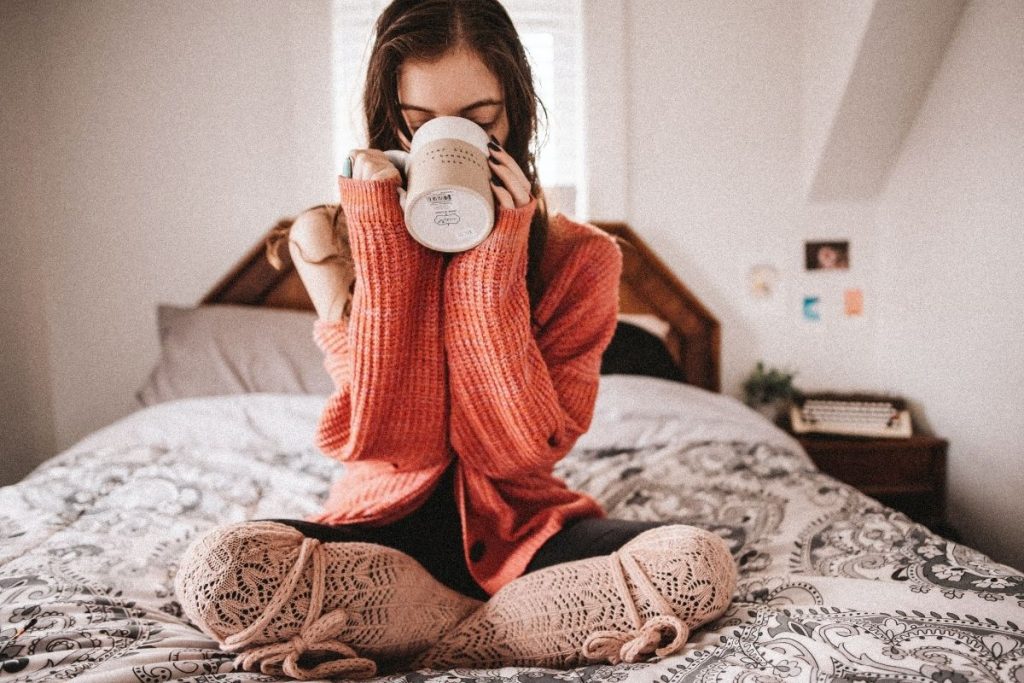 a girl with dark hair sitting alone crosslegged on her bed drinking from a coffee mug