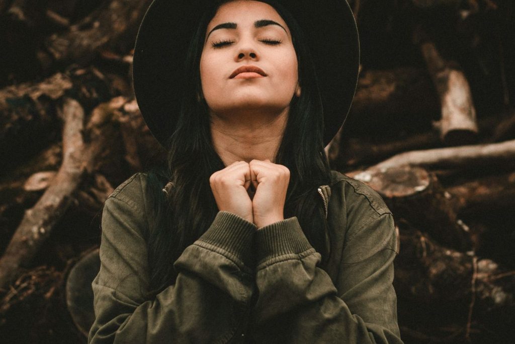 a woman with long dark hair wearing an olive jacket and a hat is holding her hands together in prayer, eyes closed and face tilted toward the sky