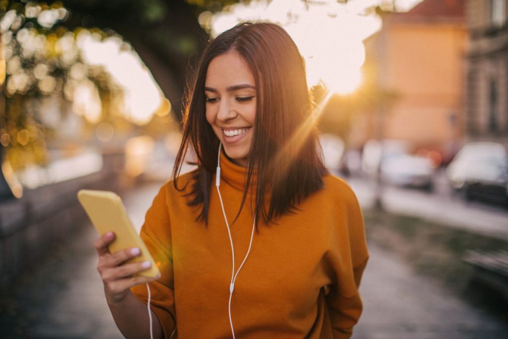 a woman with brown h air in an orange sweater is walking outside staring at her phone and smiling, she has earbuds in.