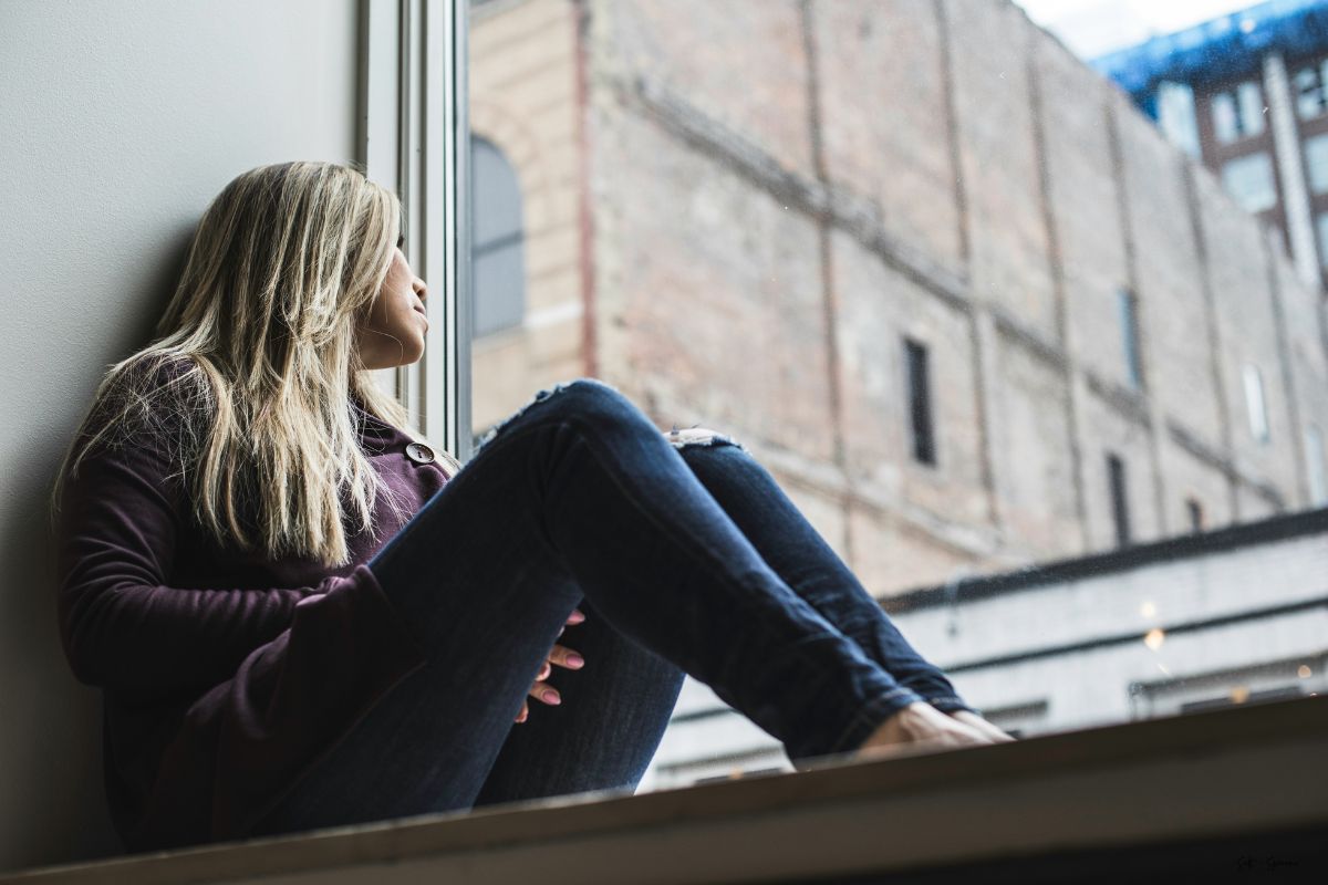 a woman sitting inside along a window ledge staring outside