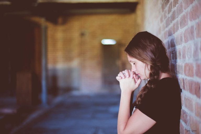 a woman with her back against a brick wall, eyes closed and hands clasped together in prayer