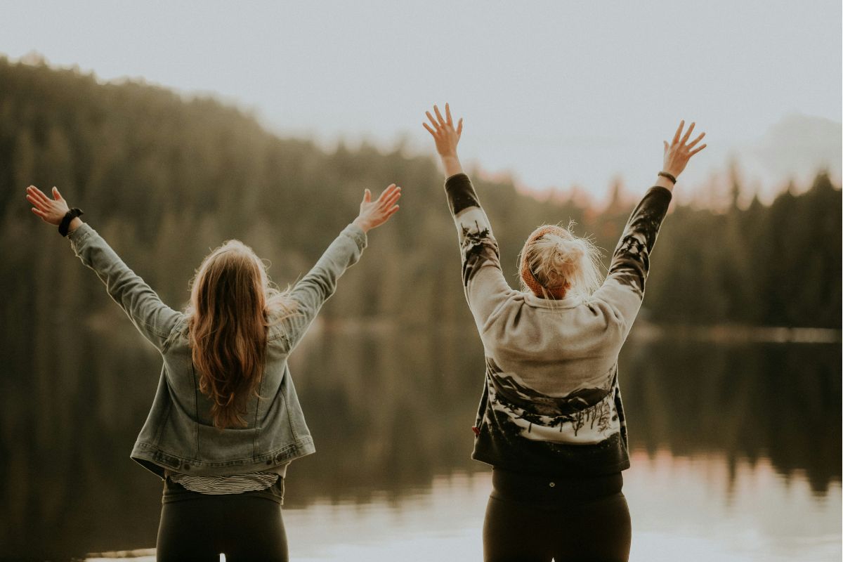 two women standing outdoors facing a lake with their arms up in the air as if praising God