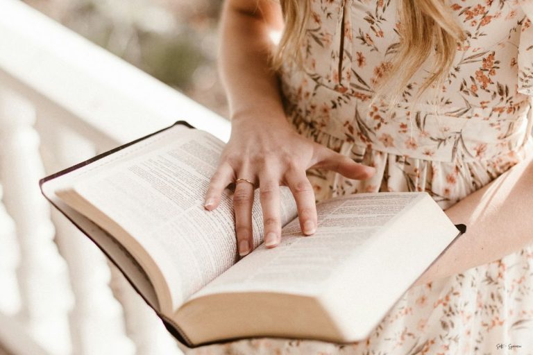 a woman in a floral dress standing and holding a bible with her hand on the pages as if to keep her place