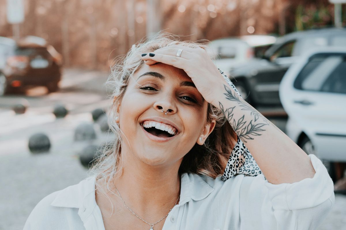 a young woman with a tattoo on her arm laughing and smiling toward the camera, you can see cars behind her